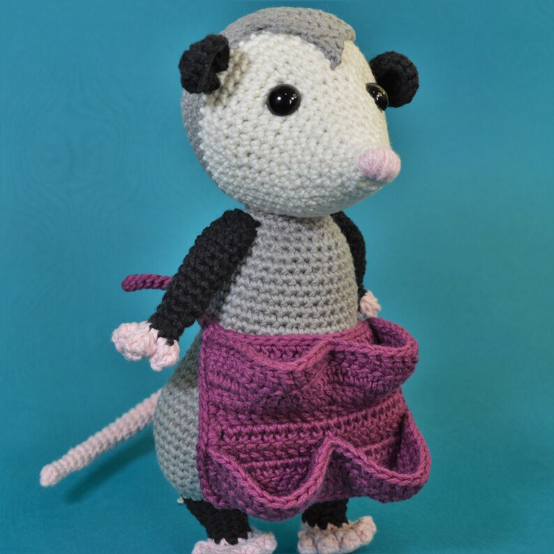 Pu&ograve; includere: Una bambola di opossum all'uncinetto che indossa un grembiule rosa con tasche. La bambola ha occhi neri, un corpo grigio e una coda nera e grigia.