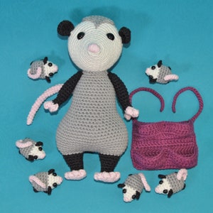 Pu&ograve; includere: Un opossum all'uncinetto grigio e bianco con un naso rosa e occhi neri. L'opossum tiene una borsa rosa all'uncinetto. Ci sono sei opossum pi&ugrave; piccoli all'uncinetto intorno all'opossum pi&ugrave; grande.