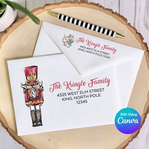 以下が含まれることがあります： 白い封筒に、赤と金のくるみ割り人形のイラストと「The Kringle Family 4325 West Elm Street King, North Pole 12345」という文字が印刷されています。封筒は木製の表面の上に置かれています。