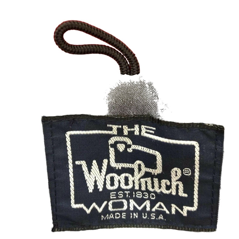May include: A navy blue fabric label with white stitching. The label reads "The Woolrich Woman Est. 1830 Made in U.S.A."