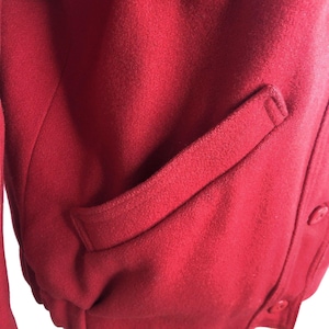 May include: A close-up of a red wool jacket with a single pocket on the right side. The jacket has a button closure.