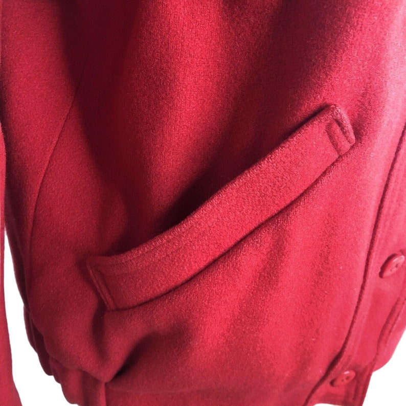 May include: A close-up of a red wool jacket with a single pocket on the right side. The jacket has a button closure.