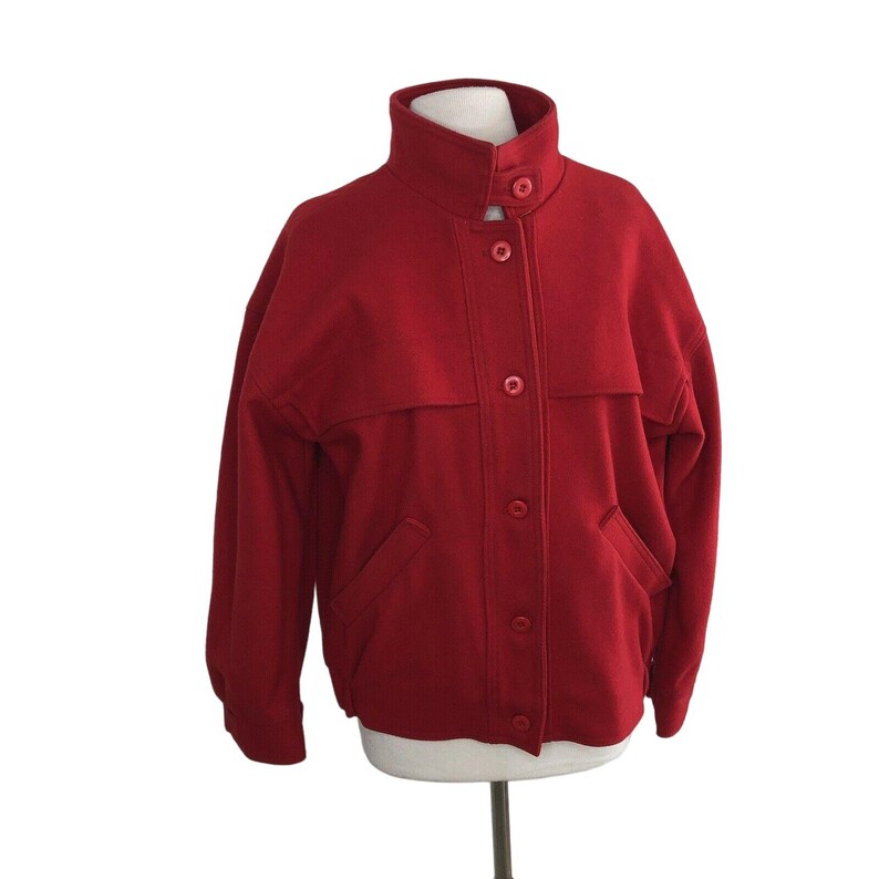 May include: A red wool jacket with a stand-up collar and button closure. The jacket has two flap pockets on the front and a single button on each cuff.