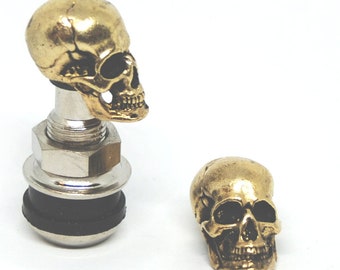 May include: Two gold-colored skull tire valve stem caps. The cap on the left is attached to a silver valve stem. The cap on the right is separate.