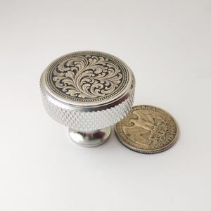 May include: A silver-colored knob with a detailed floral design on top. The knob has a textured, ridged edge and a small, round base. A US quarter coin is next to the knob for size comparison.
