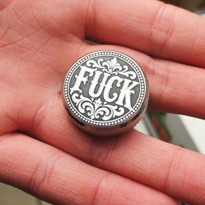 Puede incluir: Un objeto metálico circular con acabado gris oscuro que presenta la palabra FUCK grabada en una fuente estilizada y llamativa. El texto está rodeado por intrincados adornos florales simétricos y un borde decorativo de cuentas.