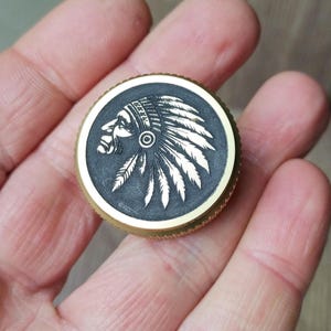 Könnte beinhalten: Ein rundes, messingfarbenes Objekt mit einer detaillierten Gravur eines indianischen Häuptlings im Profil. Der Häuptling trägt einen Federkopfschmuck. Der Hintergrund ist dunkel und der Rand hat eine strukturierte Kante.