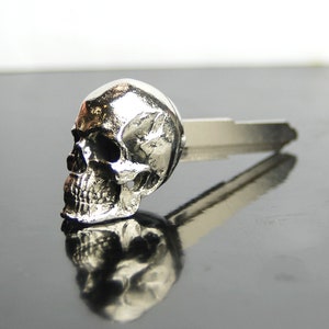 以下が含まれることがあります： 詳細な頭蓋骨のデザインが施された銀製のキーホルダー。頭蓋骨は銀製のキーホルダーに取り付けられています。