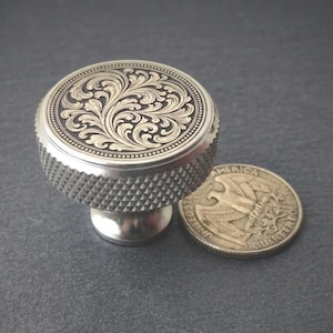 Può includere: Un pomello color argento con un design floreale inciso sulla parte superiore. Il pomello ha un lato strutturato e scanalato e una base liscia e arrotondata. Una moneta da un quarto di dollaro è accanto al pomello per il confronto delle dimensioni.