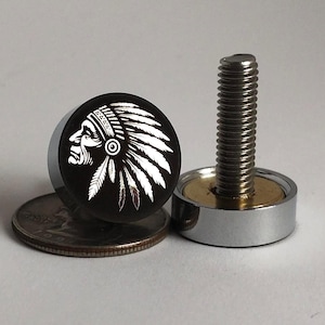 以下が含まれることがあります： ネイティブアメリカンの横顔デザインの黒とシルバーのマグネットスタッド。スタッドは丸く、白いグラフィックで頭飾りをつけた横顔が描かれています。シルバーのボルトが、シルバーとゴールドのマグネットベースに取り付けられています。