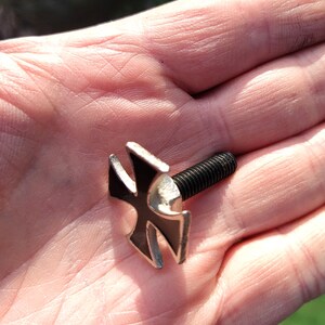 Op de afbeelding: Een zwart metalen kruis met een zilveren afwerking, bevestigd aan een schroef. Het kruis is een populair symbool van de bikercultuur.