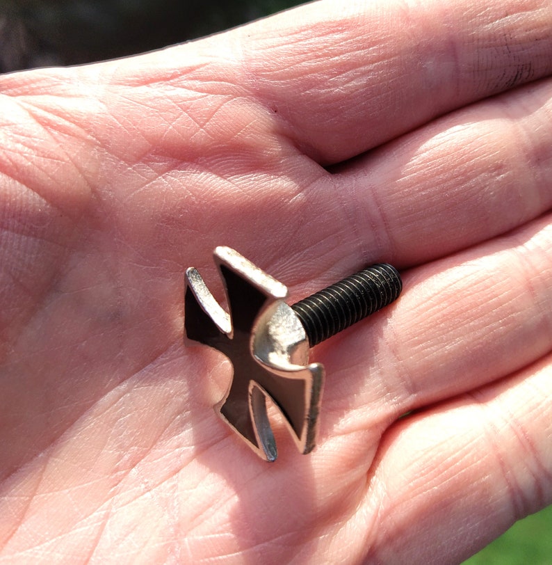 Op de afbeelding: Een zwart metalen kruis met een zilveren afwerking, bevestigd aan een schroef. Het kruis is een populair symbool van de bikercultuur.