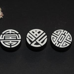 Può includere: Cinque oggetti decorativi rotondi color argento con intricati motivi bianchi, possibilmente di design asiatico. Gli oggetti sono disposti in fila su una superficie nera, con alcune rocce sullo sfondo.