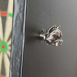 Puede incluir: Accesorio de diana con forma de cabeza de perro plateada, montado sobre una superficie negra. La cabeza del perro presenta un collar con tachuelas. El borde de una diana es visible en el fondo.