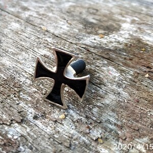 Op de afbeelding: Een zwart en zilveren metalen kruis met een schroef aan de achterkant. Het kruis ligt op een stuk verweerd hout.