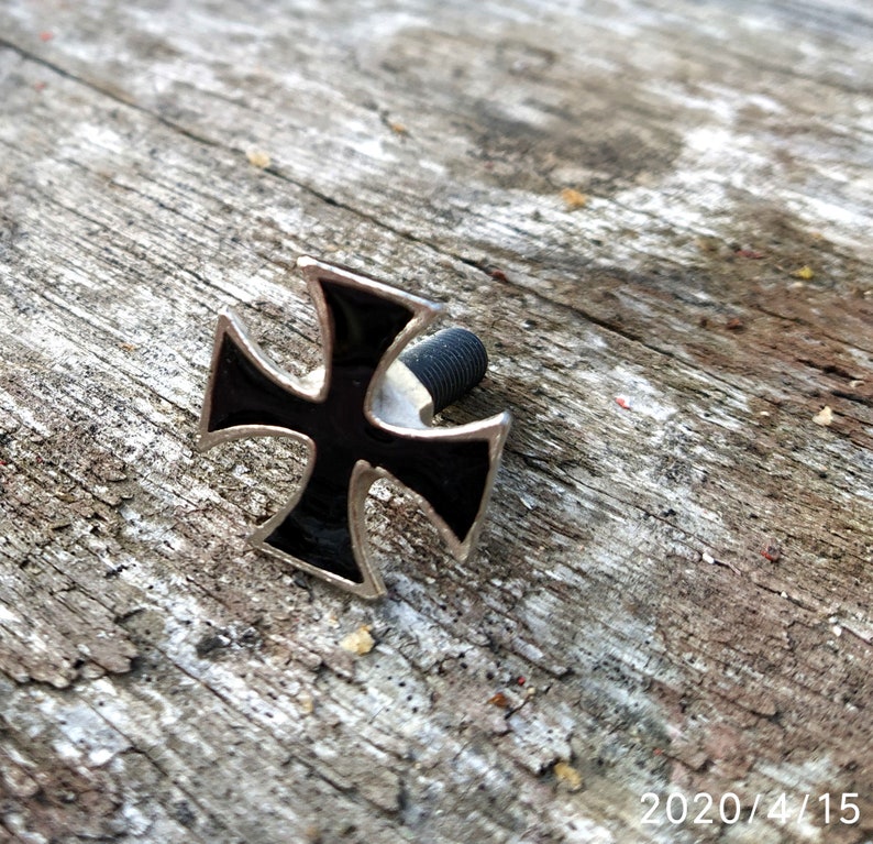 Op de afbeelding: Een zwart en zilveren metalen kruis met een schroef aan de achterkant. Het kruis ligt op een stuk verweerd hout.