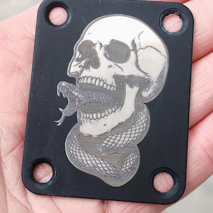 Puede incluir: Placa metálica rectangular negra con un diseño de calavera y serpiente. La calavera es blanca con sombreado detallado, y una serpiente se enrolla alrededor. La placa tiene cuatro agujeros circulares.