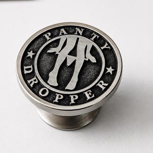 Peut inclure: Un bouton de meuble rond en métal avec une finition argentée brossée. La face présente un graphique en relief représentant des jambes et le texte PANTY DROPPER gravé dans une bordure circulaire, accentué par deux petites étoiles sur les côtés.