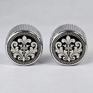 May include: A pair of circular silver-toned cufflinks featuring a textured knurled edge and a black inlay. The centre of each cufflink displays a white printed design of a fleur-de-lis topped with a small crown, surrounded by a dotted border.