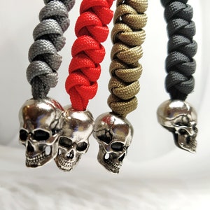 Op de afbeelding: Vier schedelvormige sleutelhangers met verschillende gekleurde paracord armbanden. De sleutelhangers zijn zilver en de armbanden zijn grijs, rood, goud en zwart.