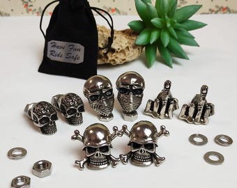 May include: A set of motorcycle accessories featuring skull and middle finger designs. The set includes a skull with a mask, a skull with a helmet, a skull with crossbones, and two middle finger designs. The accessories are made of metal and have a silver finish.