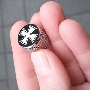 May include: A silver ring with a black face displaying a white cross design. The ring has a textured band and a round face. The ring is held in a person's hand.