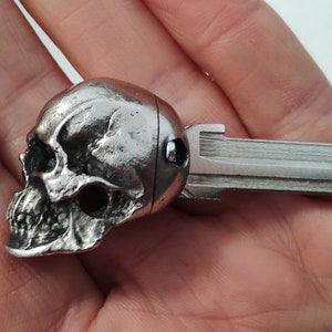 May include: A silver key featuring a skull-shaped head. The skull has detailed facial features. The key's blade is rectangular with a ridged design. The key is held in a hand.