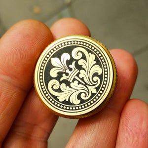 Può includere: Un disco metallico circolare color oro con un inserto centrale nero. L'inserto presenta un motivo a fleur-de-lis dorato circondato da intricati disegni a foglia. Il bordo esterno del disco ha un design testurizzato con scanalature.