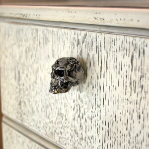 May include: A white dresser drawer with a silver skull drawer pull. The drawer is made of wood with a distressed finish.
