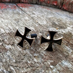 Op de afbeelding: Twee zilveren metalen kruisen met zwarte emaille centra. De kruisen liggen op een houten oppervlak.