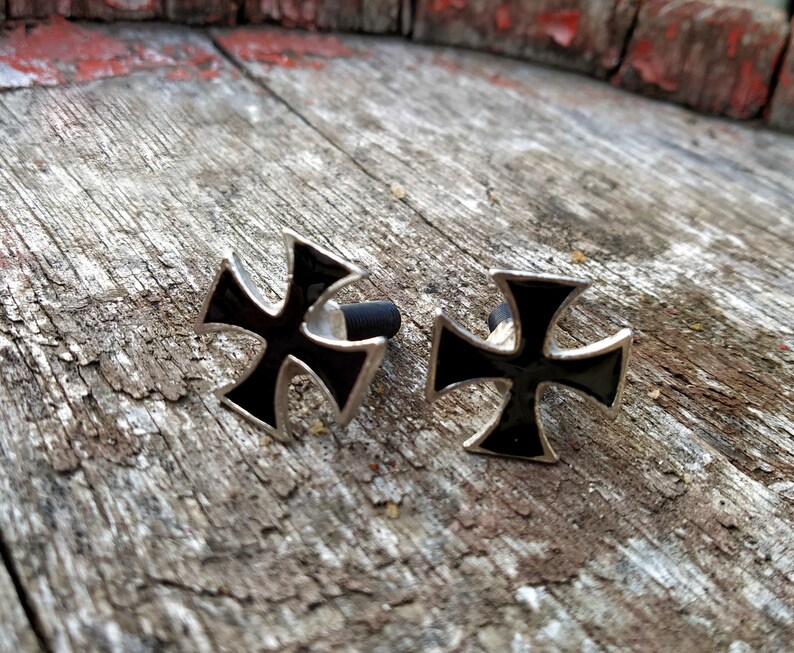 Op de afbeelding: Twee zilveren metalen kruisen met zwarte emaille centra. De kruisen liggen op een houten oppervlak.