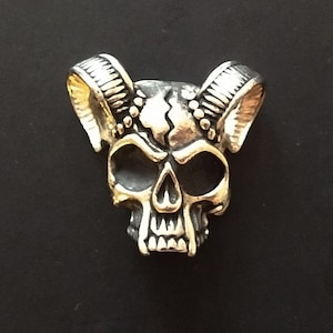 Könnte beinhalten: Ein silberner Totenkopf-Anhänger mit Hörnern, der Totenkopf hat ein rissiges Design und ein detailliertes Gesicht.