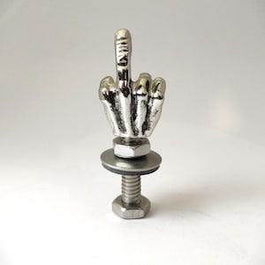 May include: A silver-coloured metal sculpture of a hand giving the middle finger, mounted on a bolt with a washer and nut. The hand is detailed with visible knuckles and lines. The sculpture is set against a plain white background.