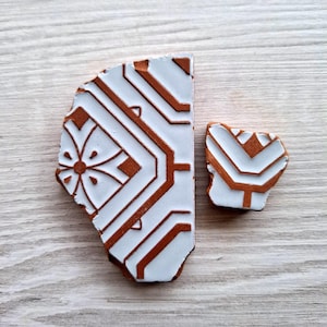 Pode incluir: Duas peças quebradas de um azulejo de cerâmica branco com um padrão geométrico castanho. O azulejo tem um fundo branco com linhas e formas castanhas.