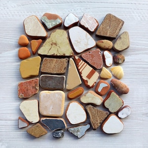 Pode incluir: Uma coleção de pequenos azulejos de cerâmica coloridos e texturizados dispostos em um padrão solto e orgânico sobre uma superfície de madeira branca.
