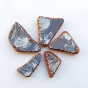 Puede incluir: Cinco piezas de vidrio marino de forma triangular irregular con bordes suaves y redondeados. Cada pieza presenta una base de color gris oscuro opaco con patrones blancos veteados y arremolinados y un borde marrón rojizo.