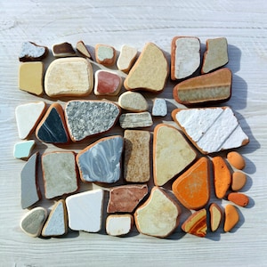 May include: A collection of colorful, irregular shaped ceramic tiles in various shades of brown, white, gray, and orange. The tiles are arranged in a random pattern on a light wood surface.