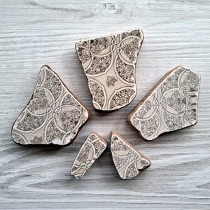Pode incluir: Cinco fragmentos cerâmicos irregulares com uma base de cor creme e um padrão repetitivo de linhas pretas com motivos circulares entrelaçados e detalhes florais. As peças variam em tamanho, com três fragmentos maiores e dois pequenos segmentos triangulares.