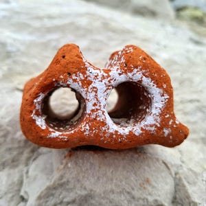 May include: A terracotta-colored brick with two holes, painted with white accents. The brick has a rough, textured surface.