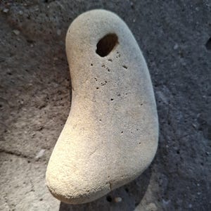 Peut inclure: Une pierre lisse de couleur gris clair, de forme arrondie, percée d'un trou près du sommet. La pierre présente une texture naturelle et patinée, posée sur une surface gris foncé. La forme de la pierre est organique et unique.