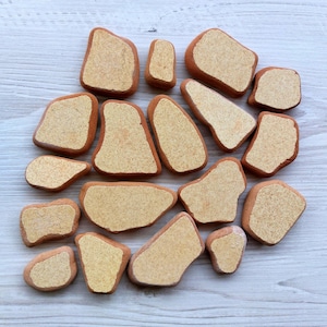 Könnte beinhalten: Eine Sammlung von 15 unregelmäßig geformten, hellbraunen Keramikfliesen mit einer gesprenkelten, beigefarbenen Oberfläche. Die Fliesen sind in einer Gruppe auf einem weißen Holzgrund angeordnet.