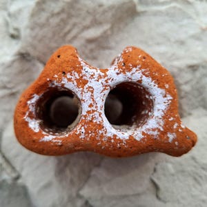 May include: A terracotta-colored ceramic object with two holes, resembling a small cave, covered in white paint splatter.