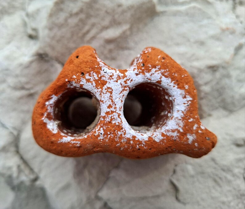 May include: A terracotta-colored ceramic object with two holes, resembling a small cave, covered in white paint splatter.
