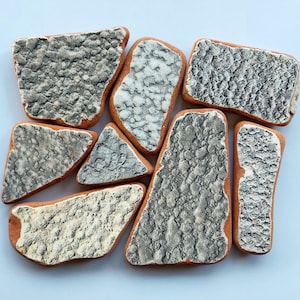 Pode incluir: Um conjunto de oito azulejos de cerâmica com uma superfície texturizada e manchada em tons de cinza e branco. Os azulejos têm formas irregulares e um aspeto rústico e feito à mão.