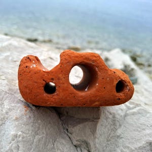 May include: A terracotta-colored ceramic object with multiple holes, resembling a small, abstract sculpture. The object is resting on a light gray, textured surface.