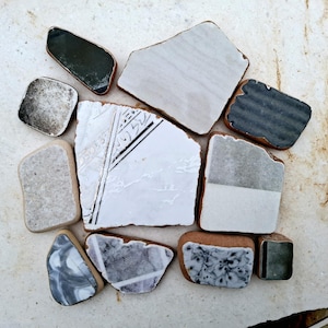 Pode incluir: Uma coleção de 10 pequenos azulejos cerâmicos de forma irregular em tons de branco, cinza e preto. Alguns azulejos têm um padrão decorativo.