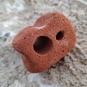 Può includere: Una presa da arrampicata a forma di roccia rossa testurizzata con due fori. La presa è progettata per pareti da arrampicata indoor.