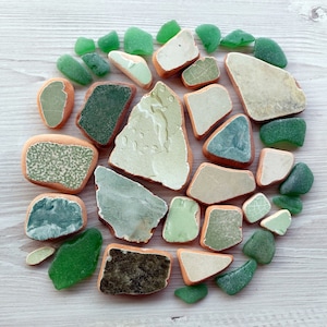 Op de afbeelding: Een verzameling van groene en witte zeeglas en keramische tegels, gerangschikt in een cirkel op een wit houten oppervlak.