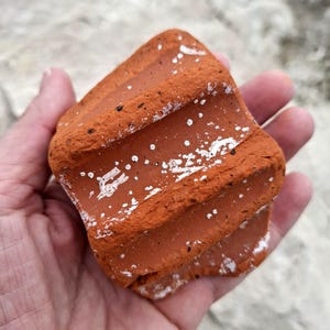 May include: A close-up of a red clay brick with white markings. The brick has a rough, textured surface and is slightly curved.