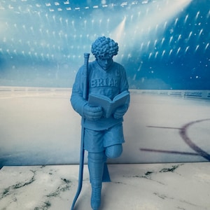Op de afbeelding: Een monochroom lichtblauw beeldje van een ijshockeyspeler die een shirt draagt met het woord BRIAR op de borst. De speler heeft krullend haar, houdt een ijshockeystick in één hand en leest een open boek dat in beide handen wordt gehouden.